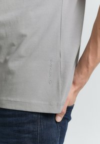 Camiseta gris claro hecha de tela suave, con un logo sutil en el lado. Tiene un corte clásico y mangas cortas, combinada con jeans oscuros.