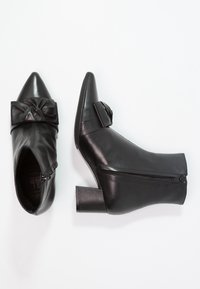 Par de botas de piel negra hasta el tobillo con cremallera lateral, tacón bloque, punta afilada y un lazo decorativo en la parte delantera.
