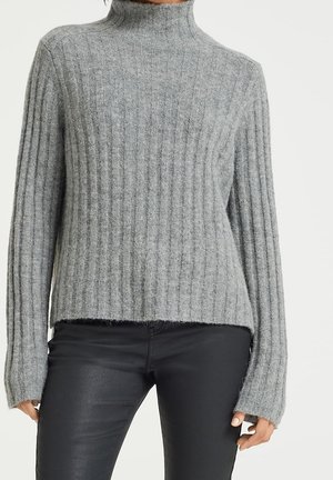 Femme portant un pull à col roulé gris côtelé et un pantalon noir ajusté, debout, les bras détendus le long du corps.