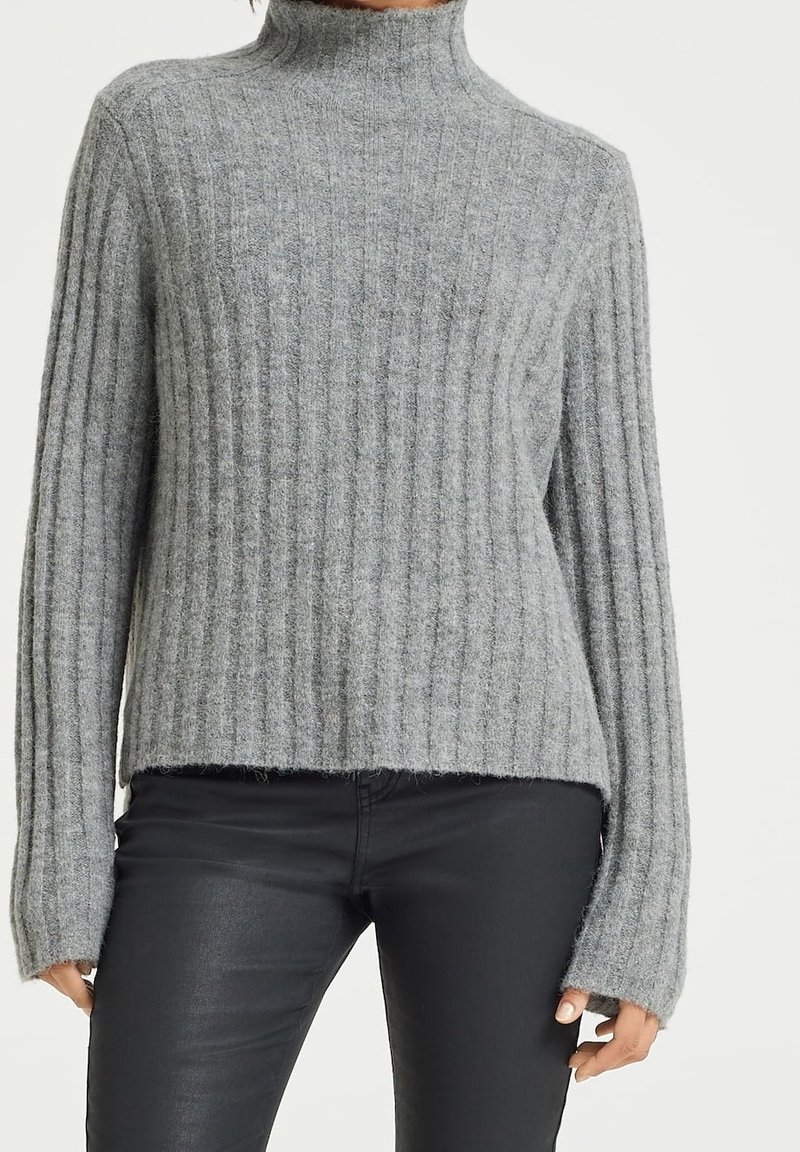 Femme portant un pull à col roulé gris côtelé et un pantalon noir ajusté, debout, les bras détendus le long du corps.