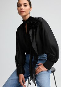 Sort bluse med flæser ved kraven og bindedetalje, lange ballonærmer, parret med høj talje blå jeans; blød tekstur, elegant design.