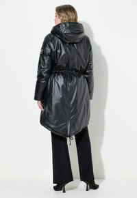 Schwarze, lange Parka mit glänzender Oberfläche, Kordelzug in der Taille und Kapuze. Bemerkenswerte Merkmale sind der asymmetrische Saum und minimale Hardwaredetails.