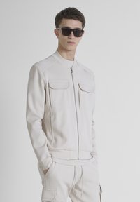 Chaqueta de cremallera de color beige claro con dos bolsillos en el pecho, puños acanalados y cuello alzado, combinada con pantalones a juego. Textura de tela suave y atlética.