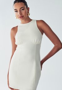 Robe blanche sans manches avec un tissu texturé, silhouette ajustée et détails plissés au niveau de la poitrine. Modèle portant de grandes boucles d'oreilles en or.