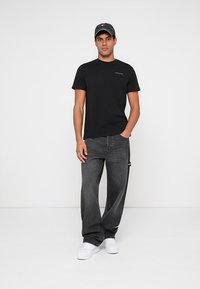 Μαύρο βαμβακερό μπλουζάκι με κοντά μανίκια και μικρό λευκό λογότυπο, συνδυασμένο με φαρδιά σκούρα γκρι τζιν και λευκά παπούτσια. Φοράει σκούρο καπέλο.