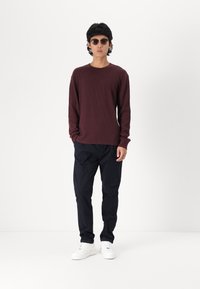 Rød langermet ribbestrikket skjorte, svarte vide bukser og hvite sneakers. Modell iført runde solbriller, står foran en ensfarget bakgrunn.