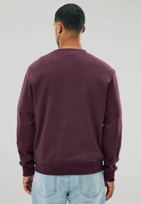 Sudadera de cuello redondo burdeos con mangas largas, puños y dobladillo acanalados, y textura suave. Combinada con jeans de mezclilla azul claro, fondo liso.