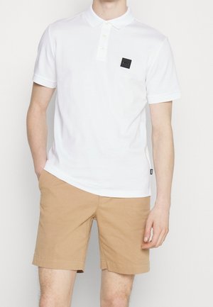 Mand iført en hvid kortærmet polo med et sort logo påsyet og beige knælange shorts, stående med den ene hånd i lommen.