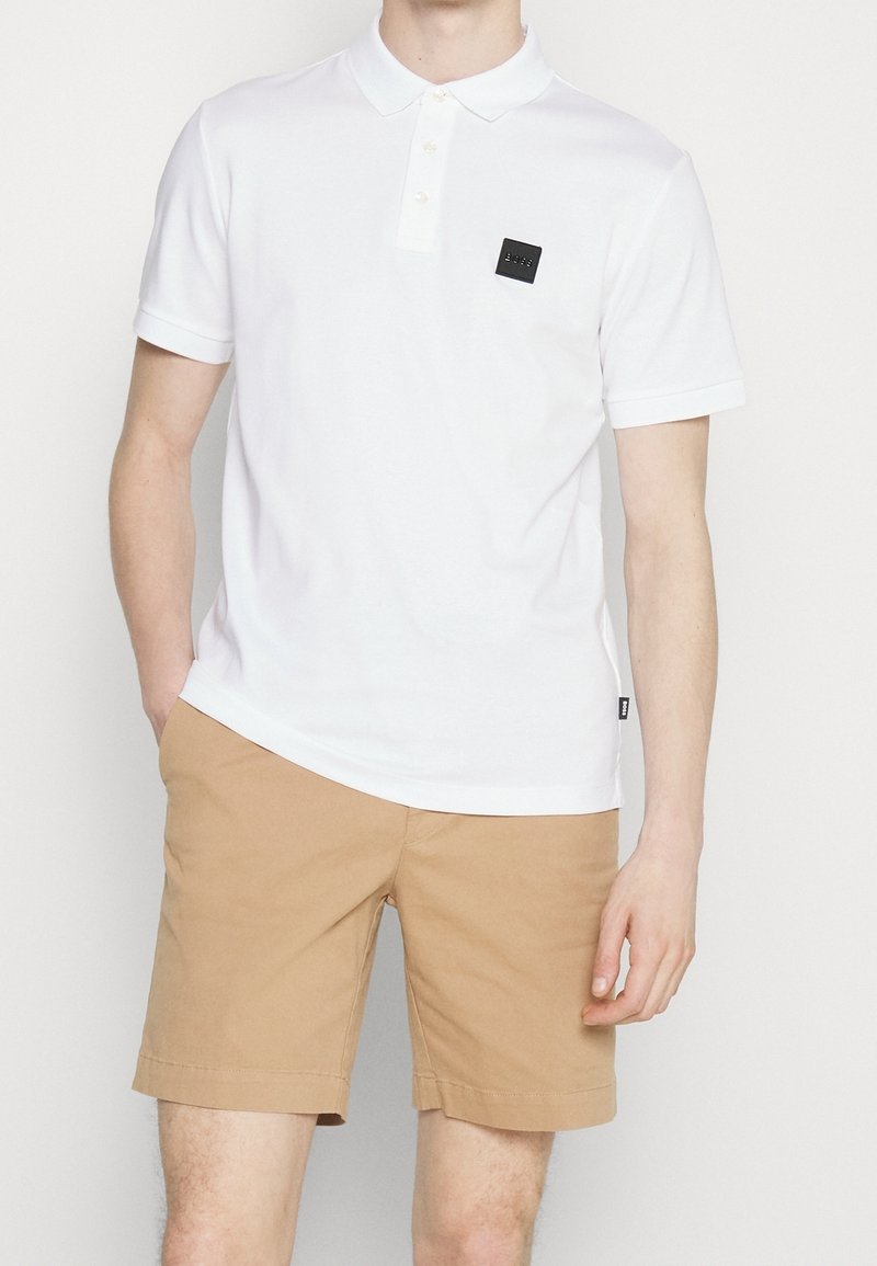 Homme portant un polo blanc à manches courtes avec un patch logo noir et un short beige jusqu'aux genoux, debout avec une main dans la poche.