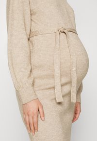 Robe en tricot beige avec une ceinture à nouer à la taille, manches longues et silhouette ajustée, conçue pour accueillir un ventre rond. Texture douce.
