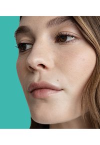 Gros plan sur un visage à la peau lisse, lèvres brun clair et yeux brun foncé. Cheveux ondulés et bruns, sur un fond turquoise lumineux.