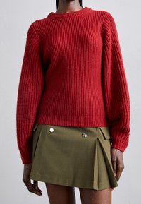 Femme portant un pull rouge côtelé à manches ballon et une mini-jupe plissée vert olive avec des boutons argentés.