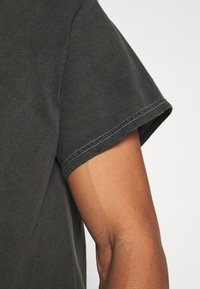 Tmavě šedé bavlněné tričko s kulatým výstřihem, krátkými rukávy a bílým prošíváním podél okraje rukávů.