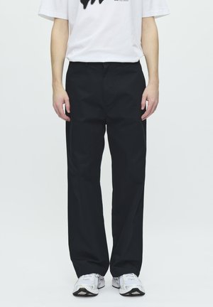 Person står iført en løs hvid T-shirt, sorte lige ben-bukser og hvide sneakers mod en ensartet lys baggrund.