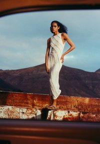 Weißes, strukturiertes Kleid mit diagonalen Mustern, knöchellang. Das Model steht auf einer rostigen Fläche unter einem wolkigen Himmel; natürliche Landschaft im Hintergrund.