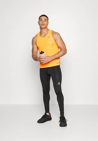 Tenue de sport pour hommes comprenant un débardeur orange vif avec un motif texturé et un legging noir ajusté. Chaussures de sport noires et bouteille d'eau incluses.