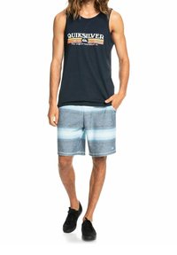 Marinblå ärmlös linne med vit och röd logotyp; blå randiga badbyxor; svarta sneakers. Avslappnad strandklar outfit med lös passform.