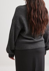 Pull tricoté gris charbon avec une coupe ample, épaules tombantes, bord côte large et tissu texturé, associé à une jupe noire à fines rayures.