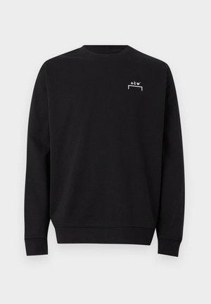 Čovjek stoji, nosi crnu majicu s malim bijelim logom, sive cargo hlače i crne cipele na jednobojnoj svijetloj pozadini.