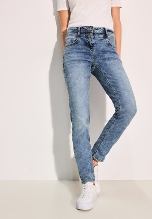 Jean en denim bleu clair avec une coupe slim, dotés d'une taille haute, de deux poches avant et d'une fermeture à bouton. Des baskets blanches complètent l'ensemble.