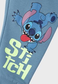 Jogging en coton bleu avec un graphique de Stitch affichant une expression espiègle et le texte "STITCH" en vert lime. Texture lisse, design décontracté.