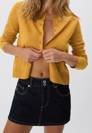 Kvinde med løst blondt hår knapper en karrygul cardigan, iført en mørk denim mininederdel med dobbelt knaplukning.