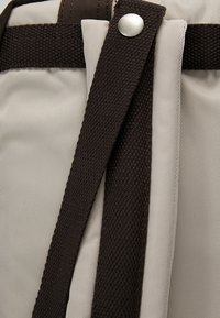 Gros plan sur un tissu beige avec des sangles sombres tissées fixées par un bouton-pression argenté.