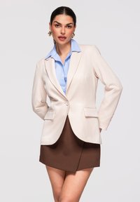 Femme aux cheveux foncés portant un blazer beige, une chemise bleu clair, une jupe marron et des boucles d'oreilles dorées, posant devant un fond gris clair.