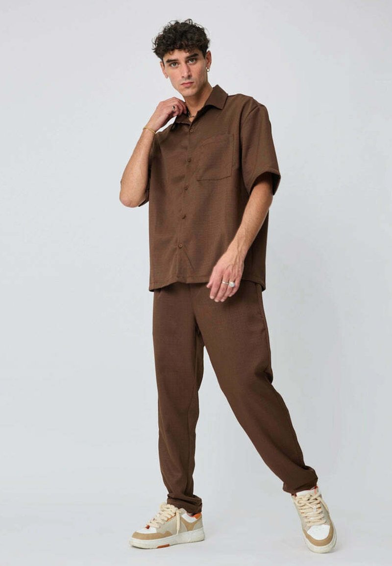 Chemise marron à manches courtes avec boutons et poche, associée à un pantalon fuselé assorti. Le mannequin porte des baskets crème et beige.