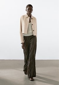 Kvinde iført en cropped beige jakke over en hvid tanktop, løse mørkebrun teksturerede bukser og sorte sandaler, stående på et ensfarvet gulv.