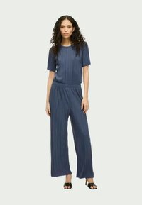 Kortsleeve ribbed-tops ja laiad püksid seljas naine lokkis juustega, kombinatsioon mustade avatud varvaste kontsatsadega.