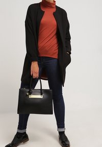 Svart lång cardigan över rostfärgad polo, mörka jeans och svarta skor. Håller en strukturerad svart handväska med guldfärgad hårdvara.