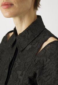Black shirt featuring a textured floral pattern, structured collar, and cut-out shoulder details. The fabric is lightweight and intricate.