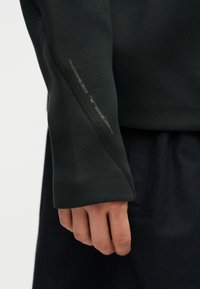 Gros plan d'une main près d'une manche longue vert foncé avec une couture diagonale visible et un texte réfléchissant "NIKE TECH" légèrement visible sur la manche.