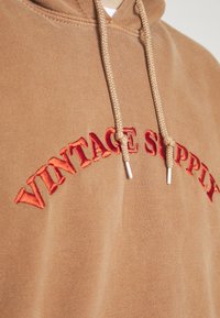 Brown hoodie with “VINTAGE SUPPLY” in red embroidery. Features beige drawstrings and a soft, textured fabric.