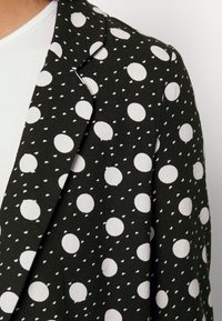 Černý blazer s bílými puntíky a malými bílými akcenty, s texturovanou tkaninou a klasickým límcem.