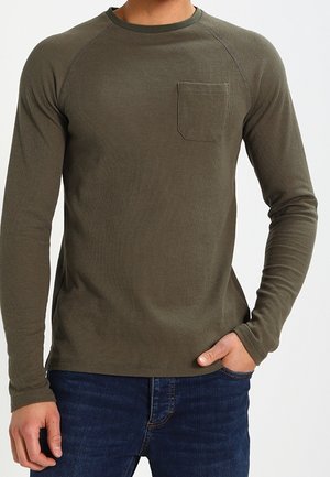 Mand iført olivengrøn langærmet skjorte med brystlomme og mørkeblå jeans, står med den ene hånd i lommen.