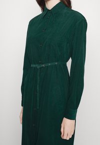 Robe chemise boutonnée vert foncé en tissu texturé, avec col, manches longues, ceinture à la taille et une poche sur le devant.