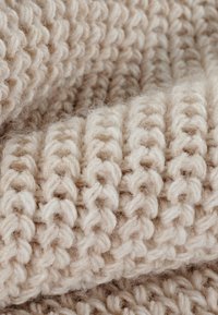 Lys beige strikket stof med et tykt, struktureret mønster. Garnet fremstår blødt og fluffy, og viser hævede sting i et gentaget design.