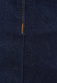 Mörkblå denimtyg med en slät textur, som har synliga sömmar i en kontrasterande gyllene färg. Närbild av materialet.