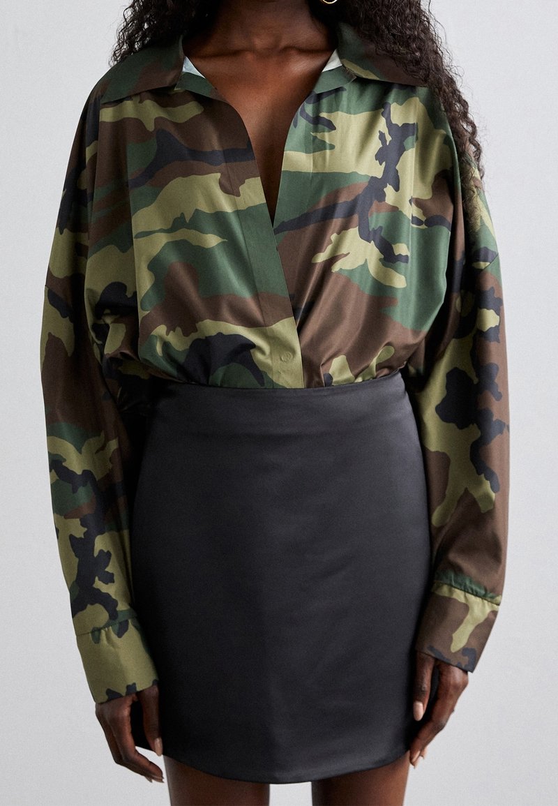 Donna che indossa una camicia larga con stampa mimetica infilata in una mini gonna nera in pelle attillata, in piedi contro uno sfondo chiaro e uniforme.