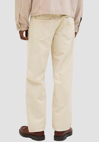 Beige Baumwollhosen mit lockerem Schnitt, ausgestattet mit zwei Gesäßtaschen, getragen mit braunen Lederschuhen und einer hellfarbigen Jacke.