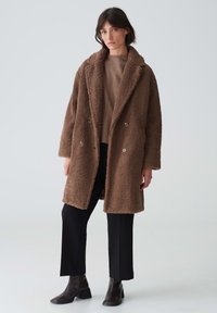 Brauner, flauschiger Wollmischmantel mit doppelter Knopfreihe, großem Kragen und zwei Knöpfen. Dazu ein beiges Sweatshirt und schwarze Hosen.