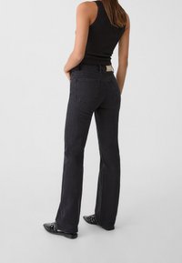 Mujer con top negro sin mangas, jeans acampanados de tiro alto en gris oscuro y zapatos negros puntiagudos, de pie contra un fondo liso.