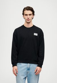 Felpa nera con scollatura a girocollo, polsini e orlo a costine. Presenta un'etichetta con logo bianco sul lato sinistro del petto. Indossata con jeans azzurri.