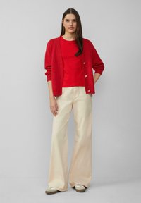 Rode cardigan over een bijpassend top met korte mouwen, gecombineerd met crèmekleurige wijde broek. De cardigan heeft een gestructureerde brei en knopen.