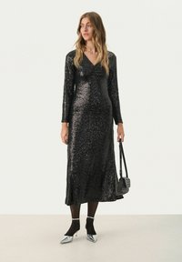 Robe midi en sequins noirs avec manches longues, décolleté en V et jupe évasée. Assortie avec des escarpins argentés à bout pointu et un petit sac noir texturé.