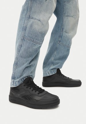 Fekete Reebok sportcipők sima bőr felsőrésszel, párnázott bokarésszel és gumitalppal, világoskék, lazán szabott farmernadrággal párosítva.