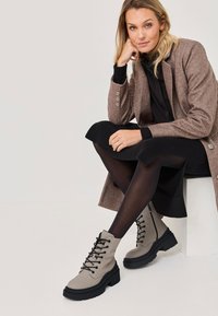 Stivaletti beige con lacci alla caviglia e suola nera spessa; realizzati in materiale simile alla pelle liscia; dettagliati con lacci neri e zip laterali.