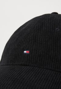 Casquette en velours côtelé noir avec une texture côtelée et un petit logo coloré avec des accents rouge, blanc et bleu sur le devant.